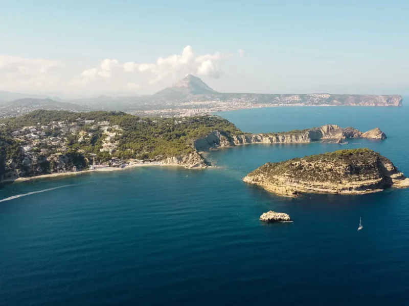 Panorámica aérea de la costa de Jávea, con colinas verdes salpicadas de residencias, una playa de arena y una isla rocosa destacada en el mar azul. Un velero y la estela de una lancha son visibles, con el Montgó al fondo.