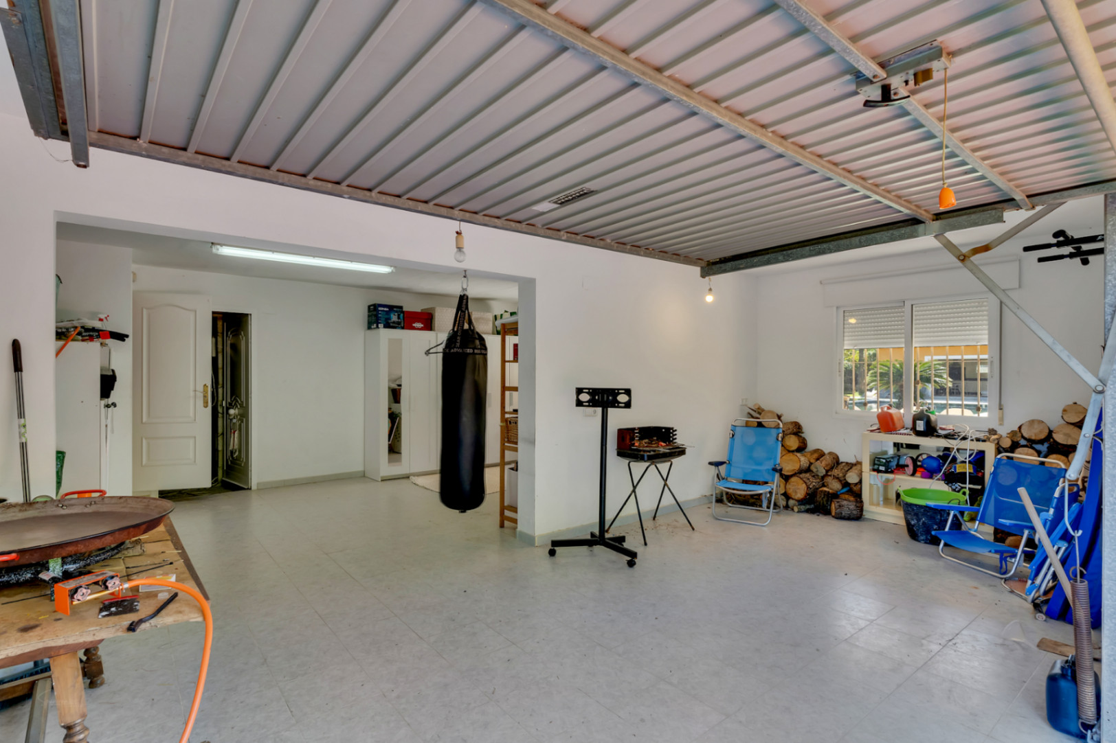Amplio garaje polivalente con techos metálicos y suelos cerámicos. Espacio versátil para almacenamiento, gimnasio y leñera con acceso directo a la vivienda.