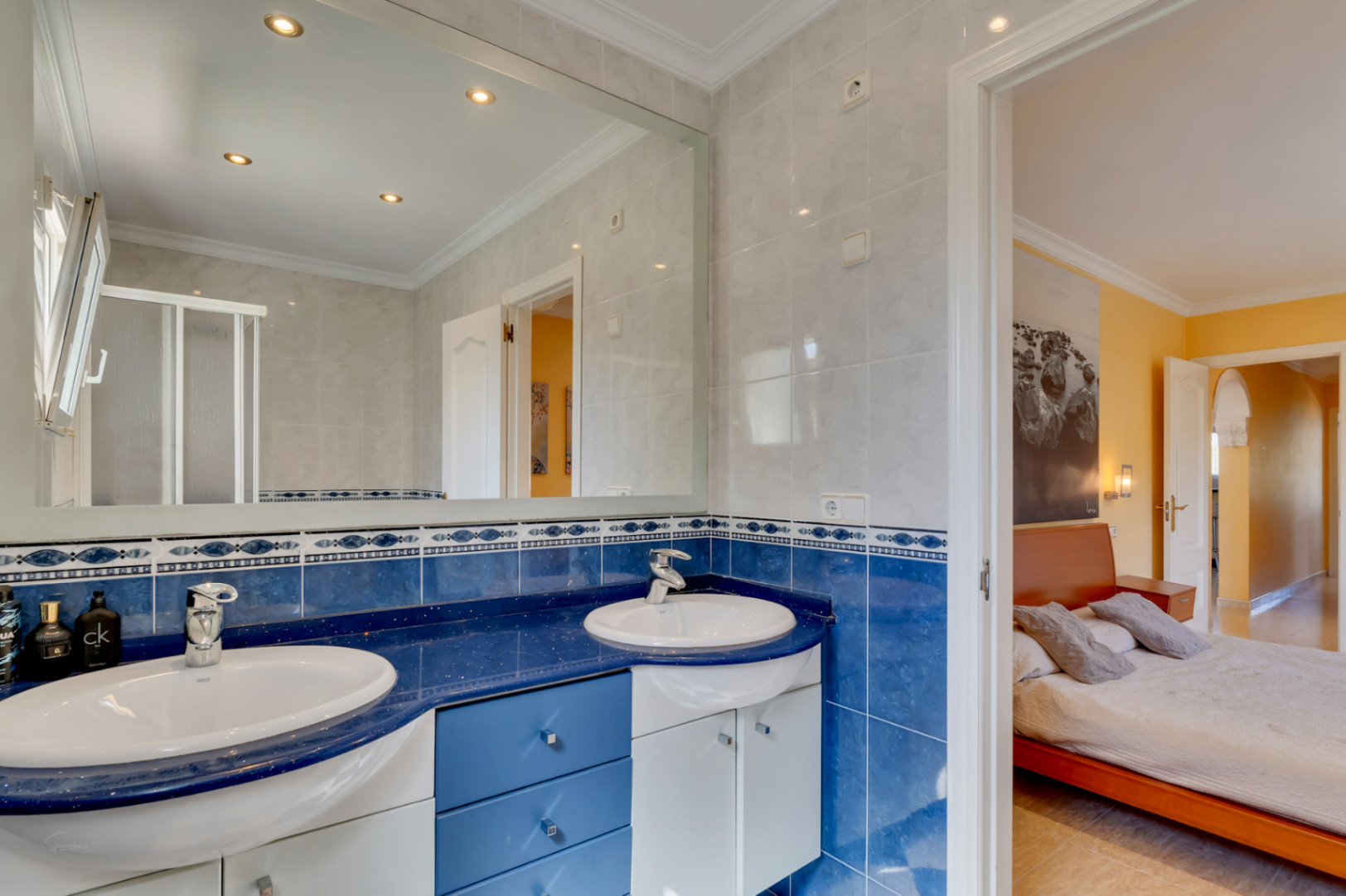 Baño con lavabo de doble seno sobre encimera azul, azulejos decorativos y gran espejo. Conexión directa con el dormitorio principal de tonos cálidos.