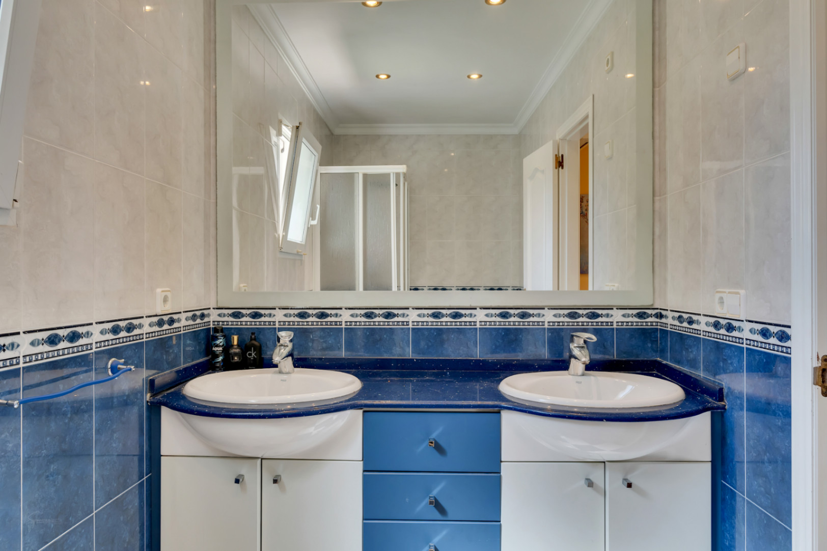 Baño con lavabo de doble seno sobre encimera azul, mueble combinado en blanco y azul, amplio espejo de pared y revestimiento cerámico con cenefa decorativa.