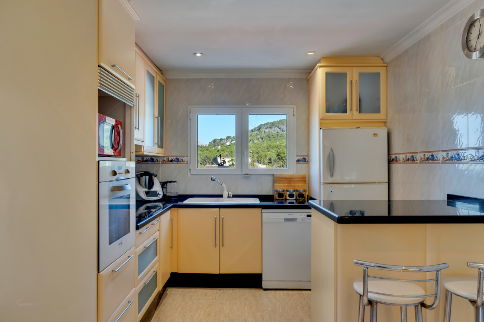 Cocina funcional con mobiliario en tono crema, encimeras de granito negro y ventana con vistas a la montaña. Incluye electrodomésticos integrados y barra de desayuno.