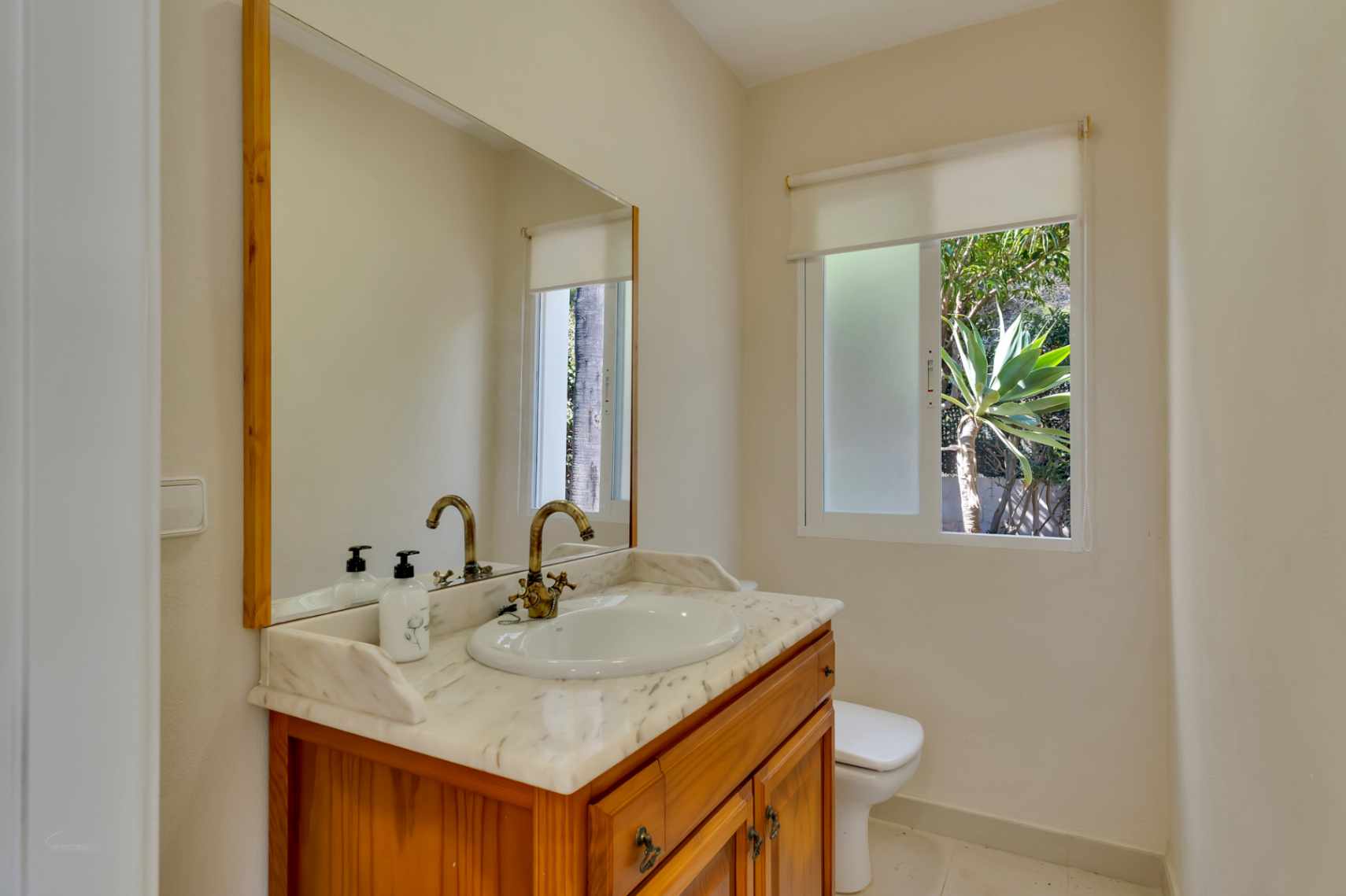 Cuarto de baño con mueble de madera, encimera de mármol y grifería de estilo clásico. Ventana exterior con vistas al jardín y abundante luz natural.