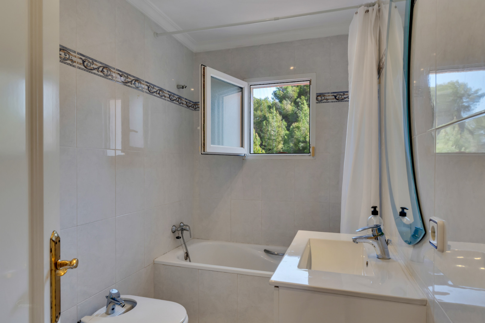Cuarto de baño funcional con bañera integrada, bidé y lavabo blanco. Incluye una ventana de PVC con vistas a la naturaleza y azulejos claros con cenefa decorativa.