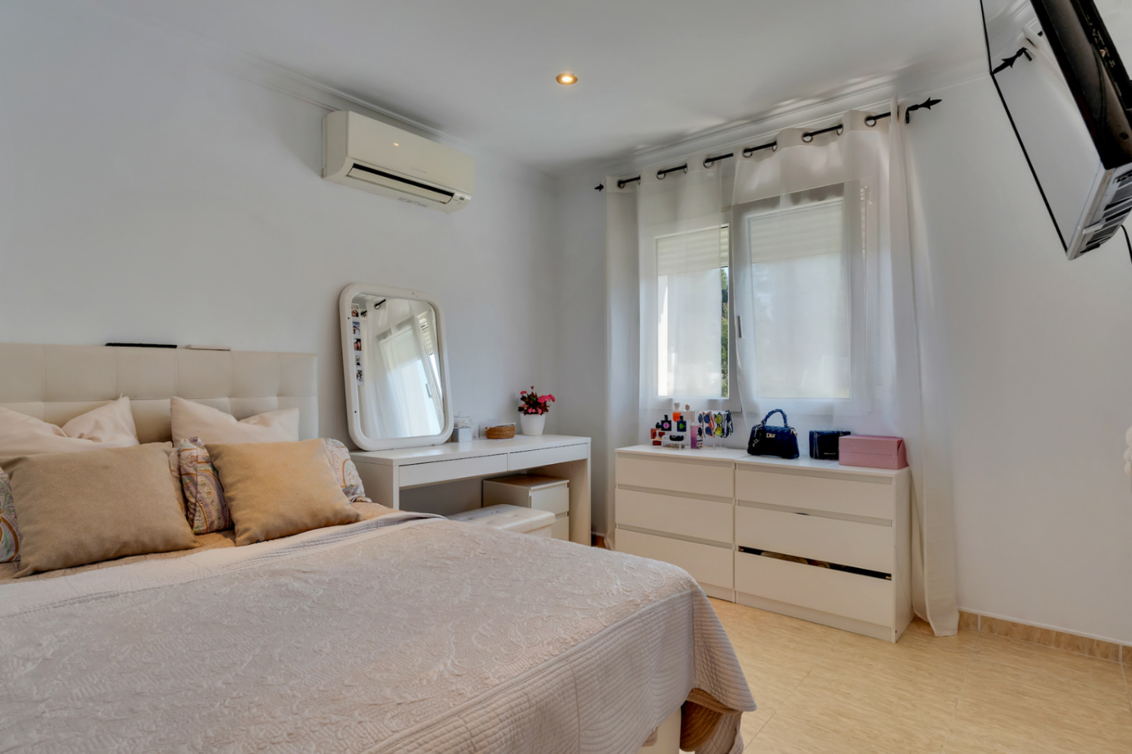 Dormitorio luminoso con mobiliario blanco, tocador con espejo, aire acondicionado y suelos cerámicos. La ventana cuenta con cortinas traslúcidas que filtran la luz natural.