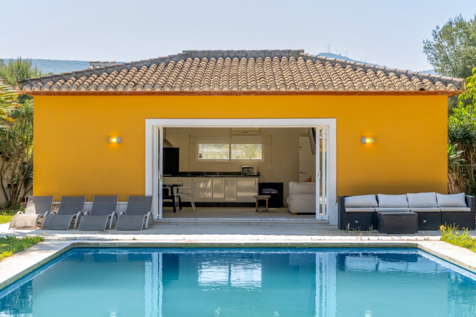 Edificio anexo junto a la piscina con cocina exterior y zona de estar, conectado mediante grandes ventanales correderos a un amplio solárium con tumbonas.