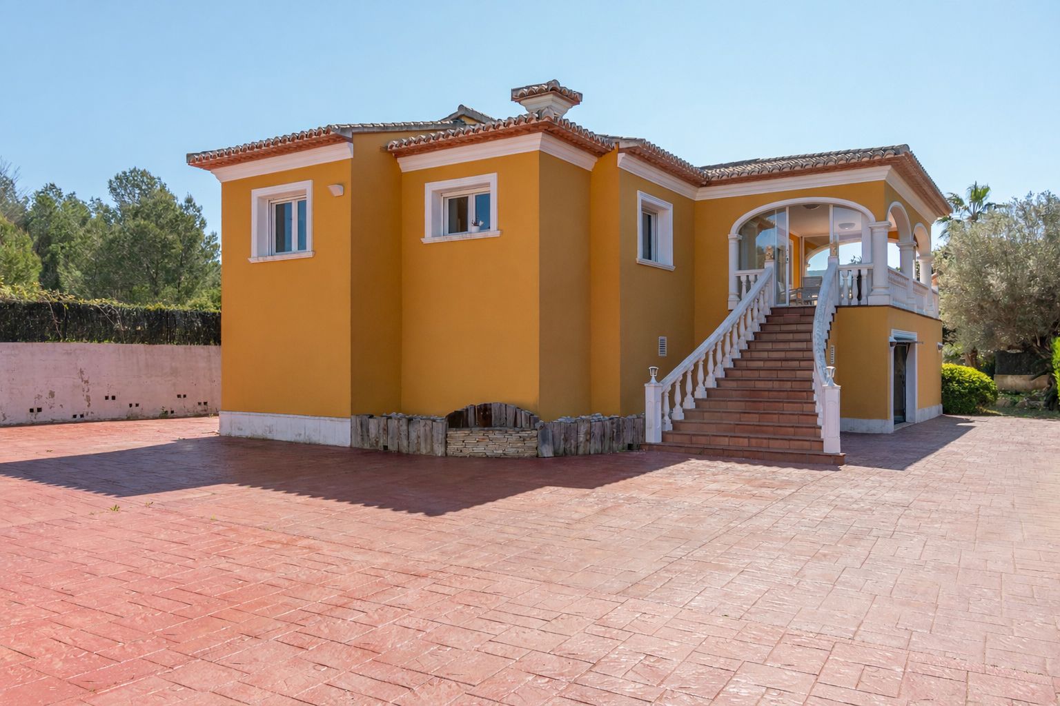 Exterior de la villa con fachada ocre, destacando una amplia zona pavimentada y una escalera señorial con balaustrada blanca que conduce a la terraza superior.