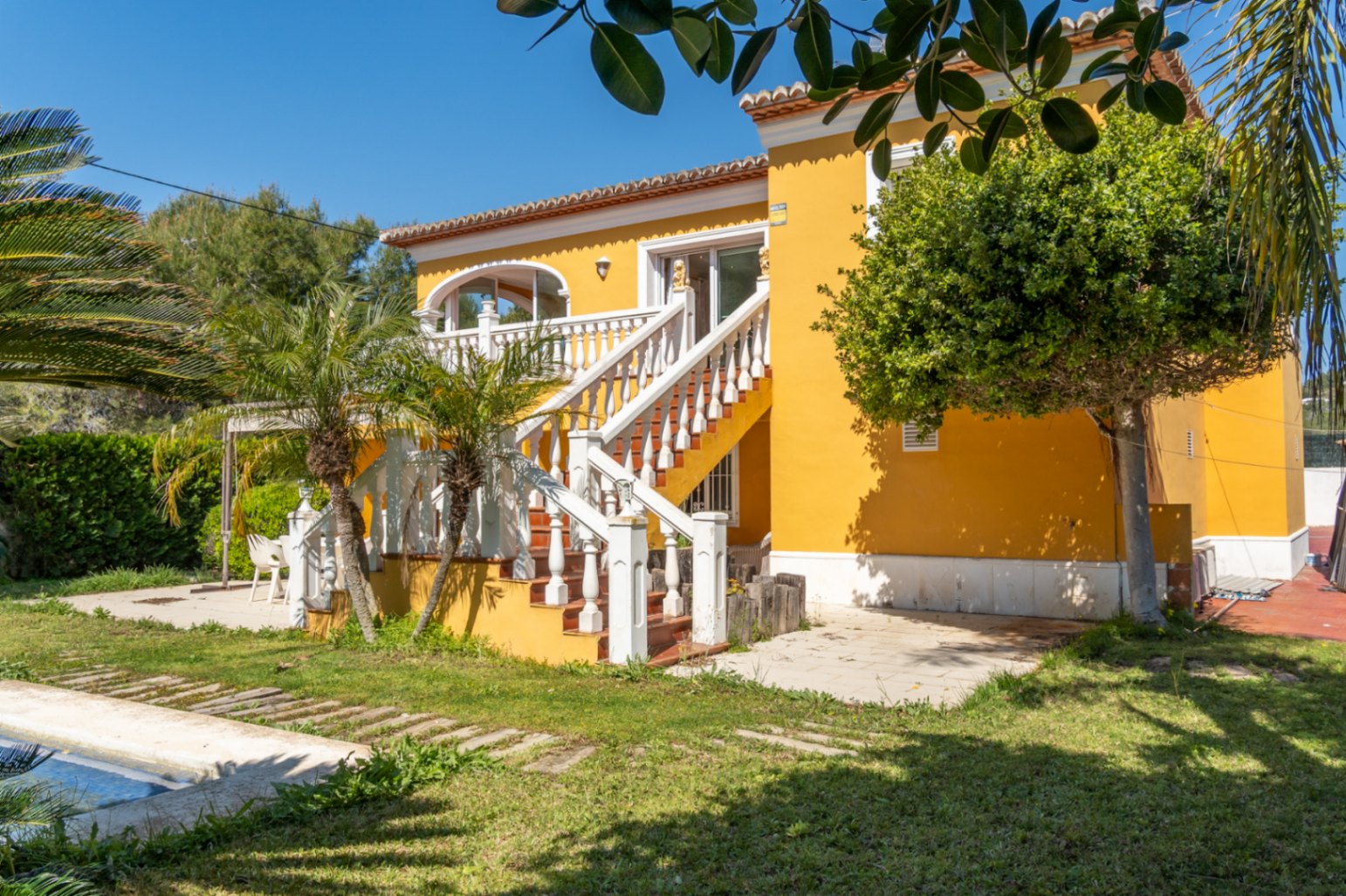 Fachada amarilla con escalera exterior de balaustrada blanca que conecta el jardín con la terraza superior. Entorno mediterráneo con palmeras y césped junto a la piscina.