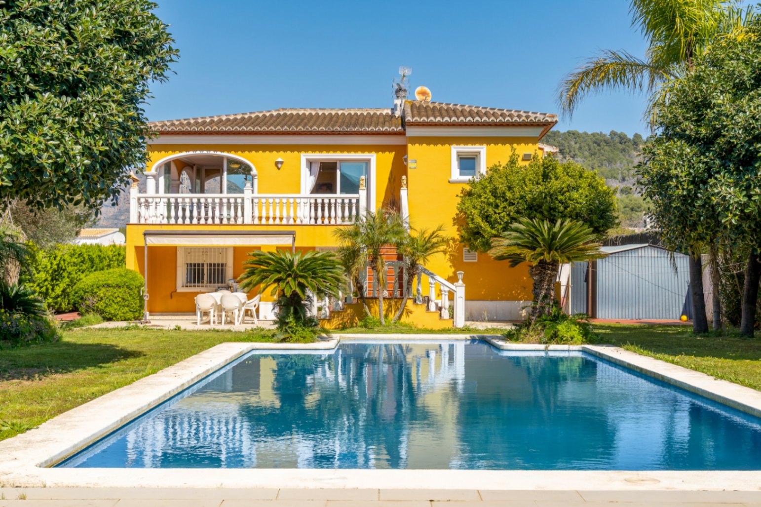 Fachada amarilla de la villa con piscina rectangular, terraza superior con balaustrada blanca y jardín mediterráneo con palmeras bajo un cielo despejado.