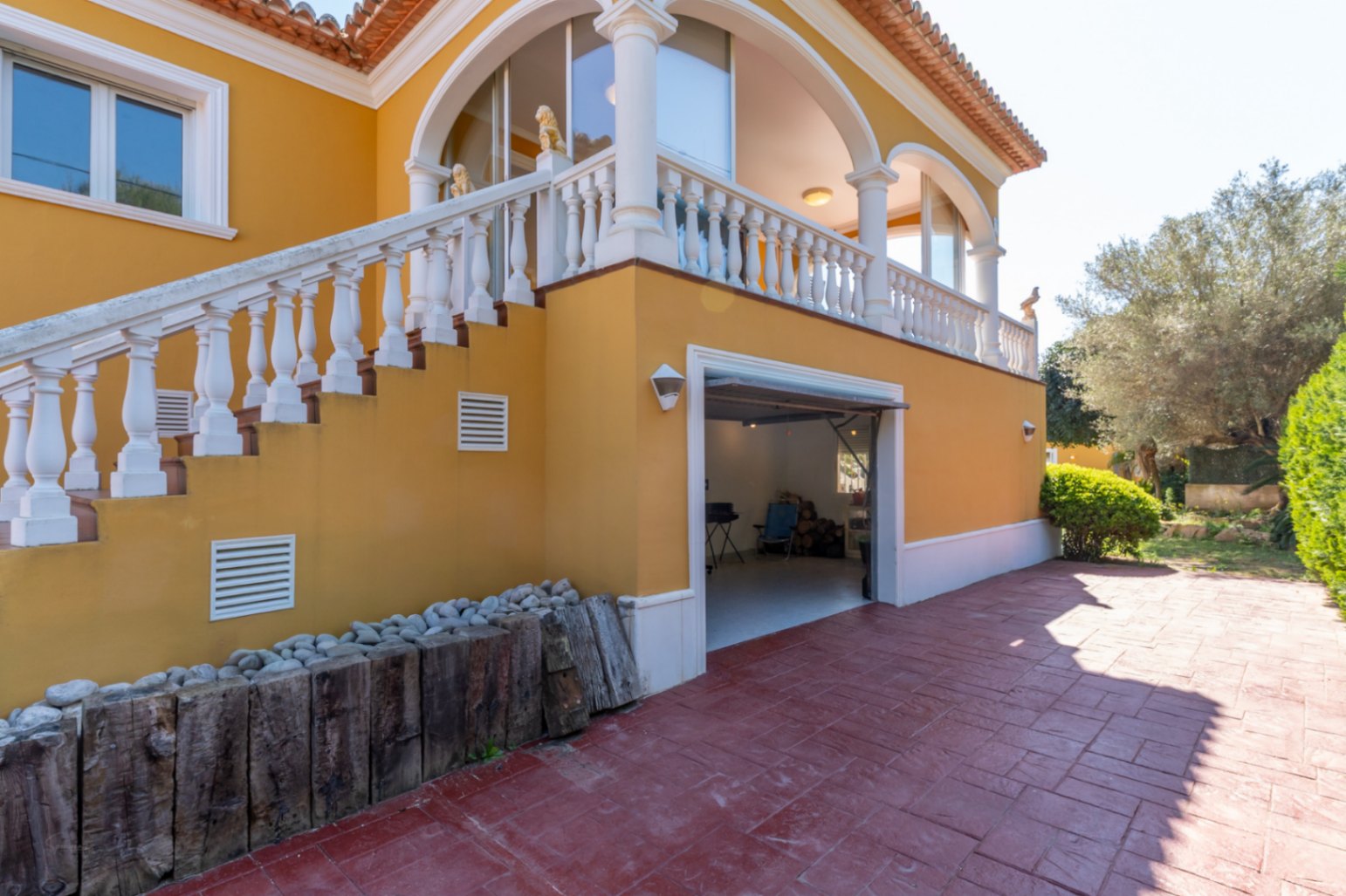 Fachada de tonos ocres con escalera exterior y balaustrada blanca que conduce a una terraza cubierta con arcos. Incluye garaje integrado y suelo de gres.