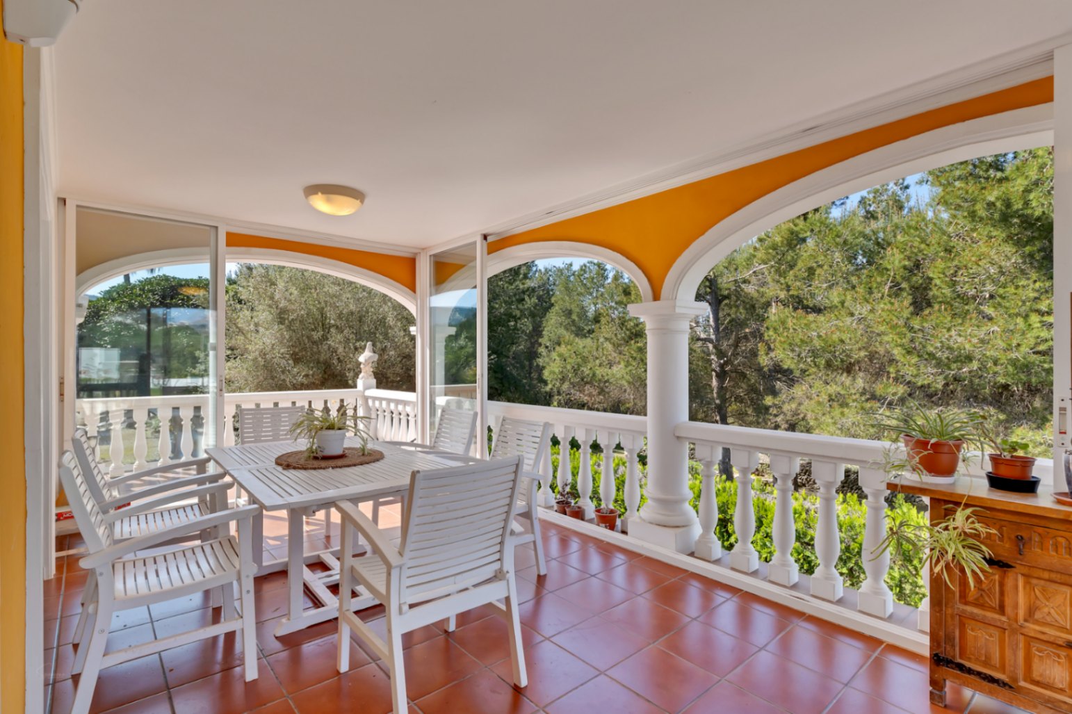 Terraza cubierta con arcos y balaustrada blanca, equipada con mobiliario de comedor de madera. Ofrece vistas despejadas a la naturaleza y cerramientos de cristal deslizantes.