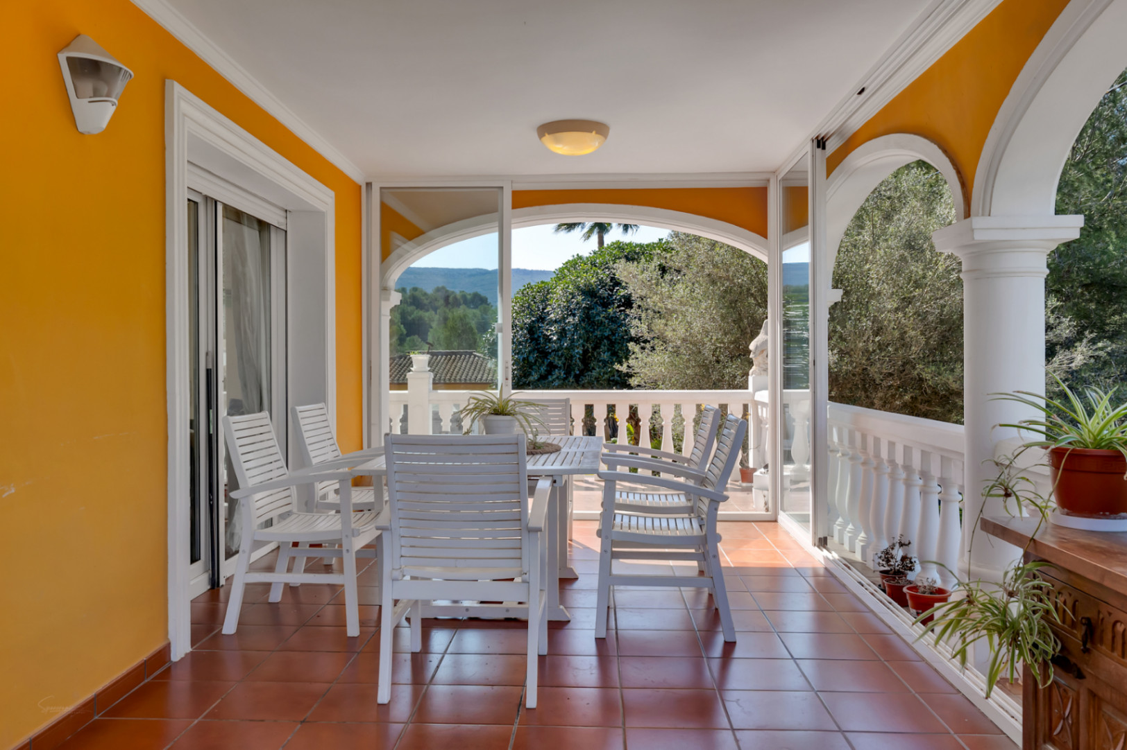 Terraza cubierta con arcos y cerramiento acristalado, equipada con mobiliario de madera blanca y suelos de gres, ofreciendo vistas despejadas a la naturaleza circundante.