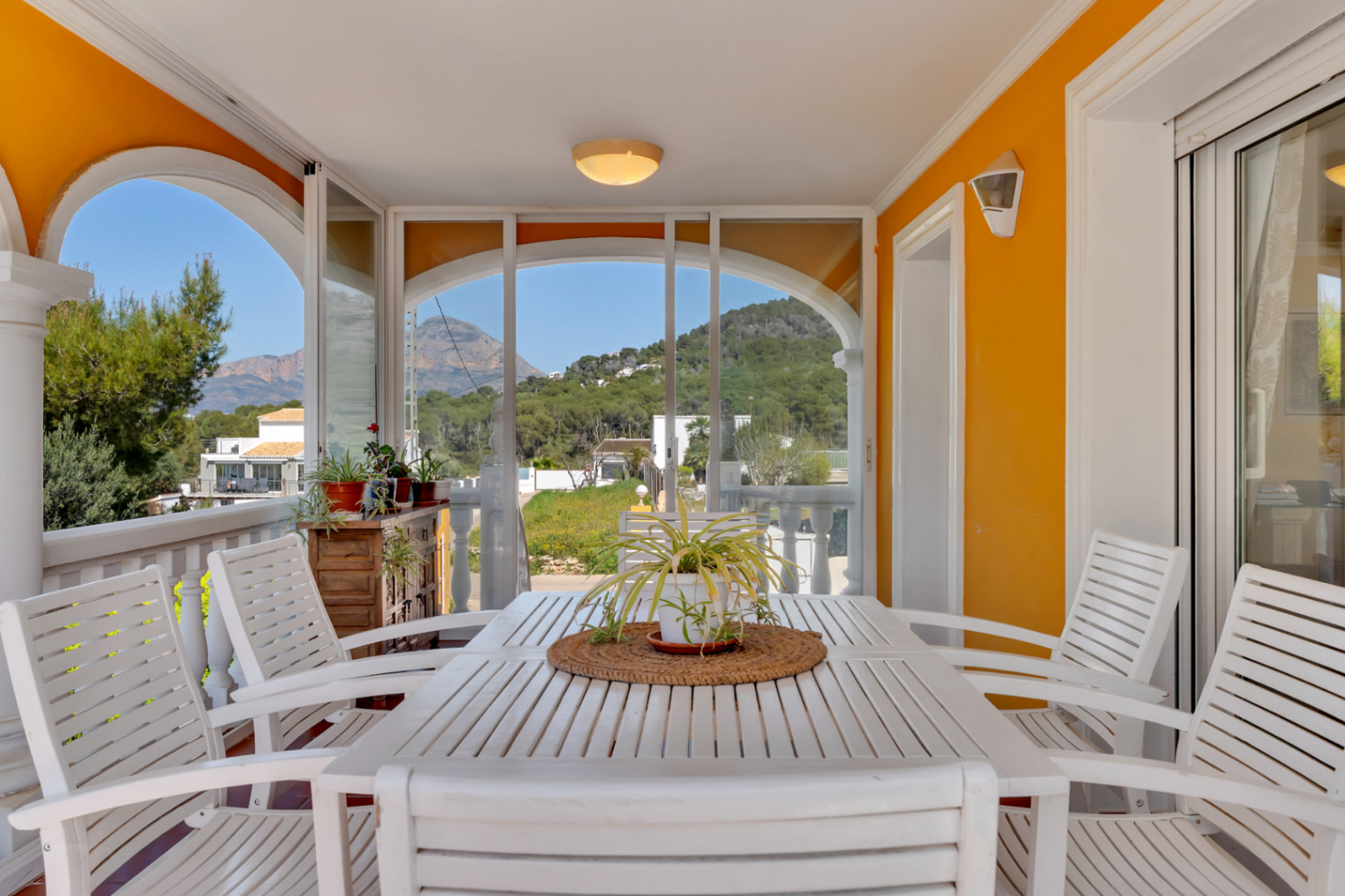 Terraza cubierta con cerramiento de cristal, mesa de madera blanca y vistas despejadas a las montañas y el entorno natural de Jávea.