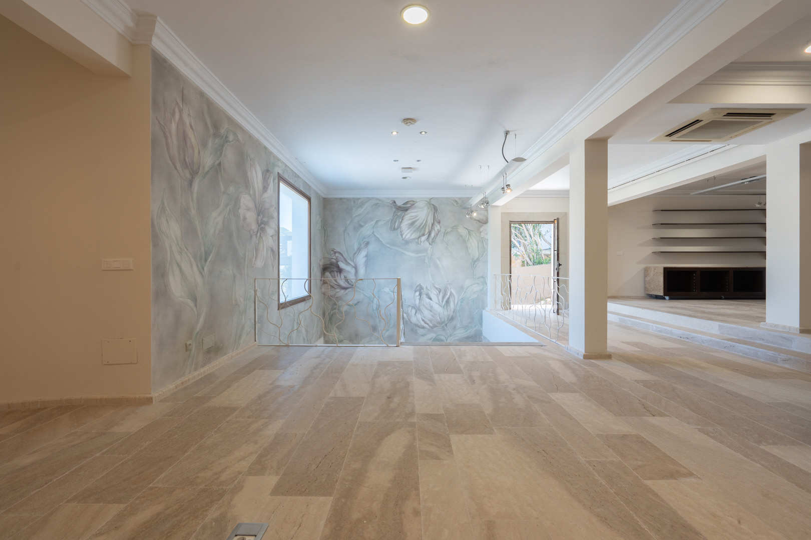 Amplio espacio diáfano con suelos de piedra natural, paredes decoradas con murales florales y barandilla de hierro forjado de diseño orgánico.