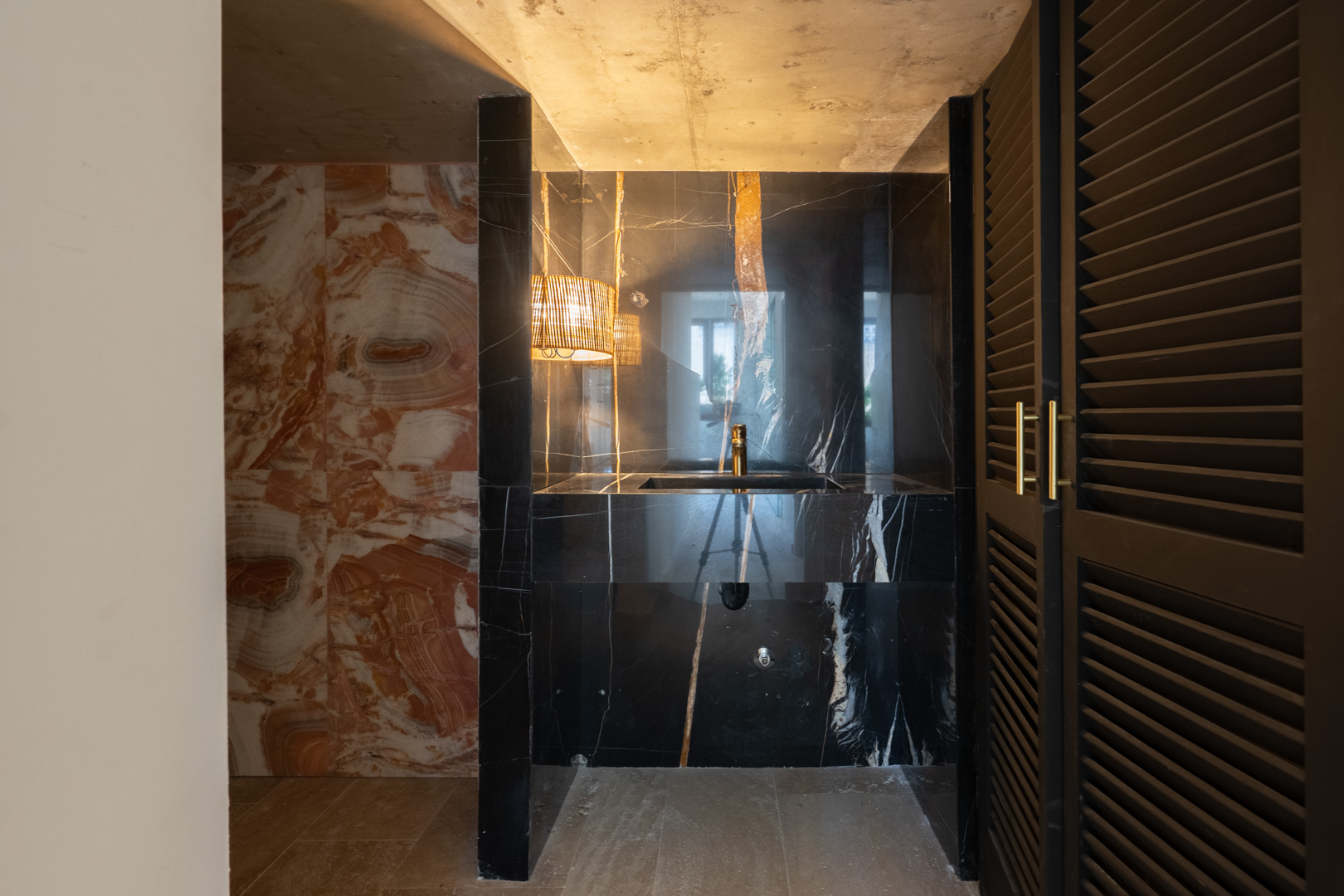Aseo de diseño con lavabo integrado en mármol negro veteado, grifería dorada, paredes de piedra natural y puertas de madera tipo persiana con herrajes dorados.