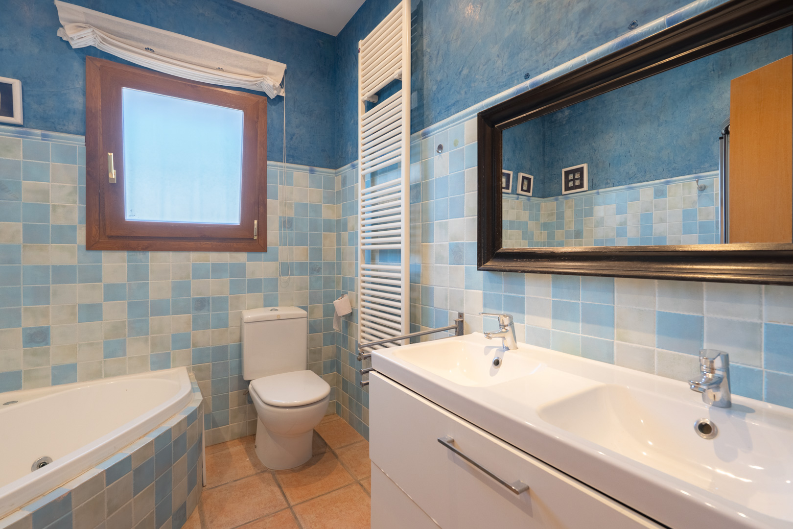 Baño con azulejos en tonos azules, lavabo doble con mueble blanco, bañera de rincón y radiador toallero sobre paredes de estuco azul.