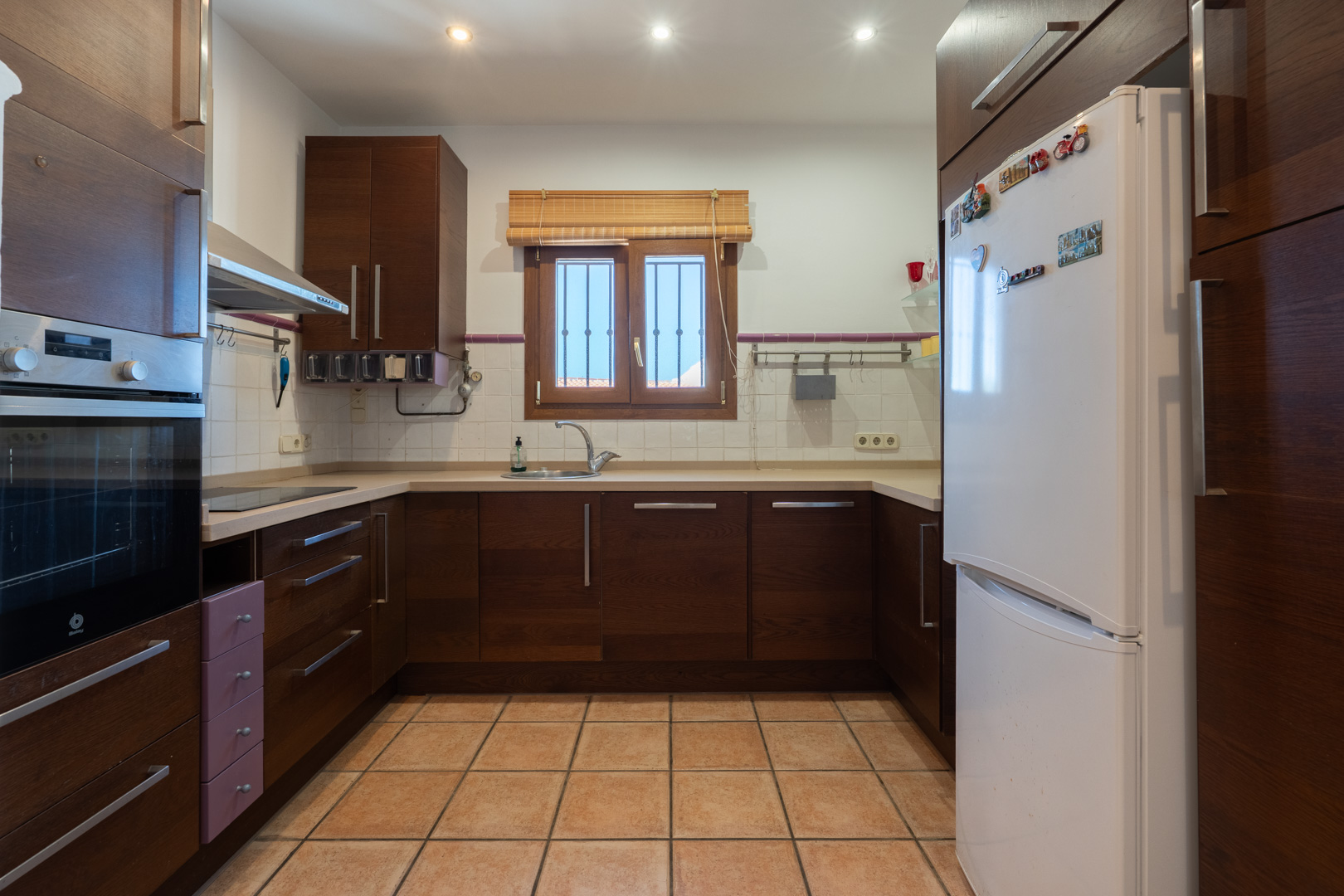 Cocina funcional con armarios de madera oscura, encimera beige y suelos de gres. Incluye horno integrado, campana extractora y ventana con persiana de madera.