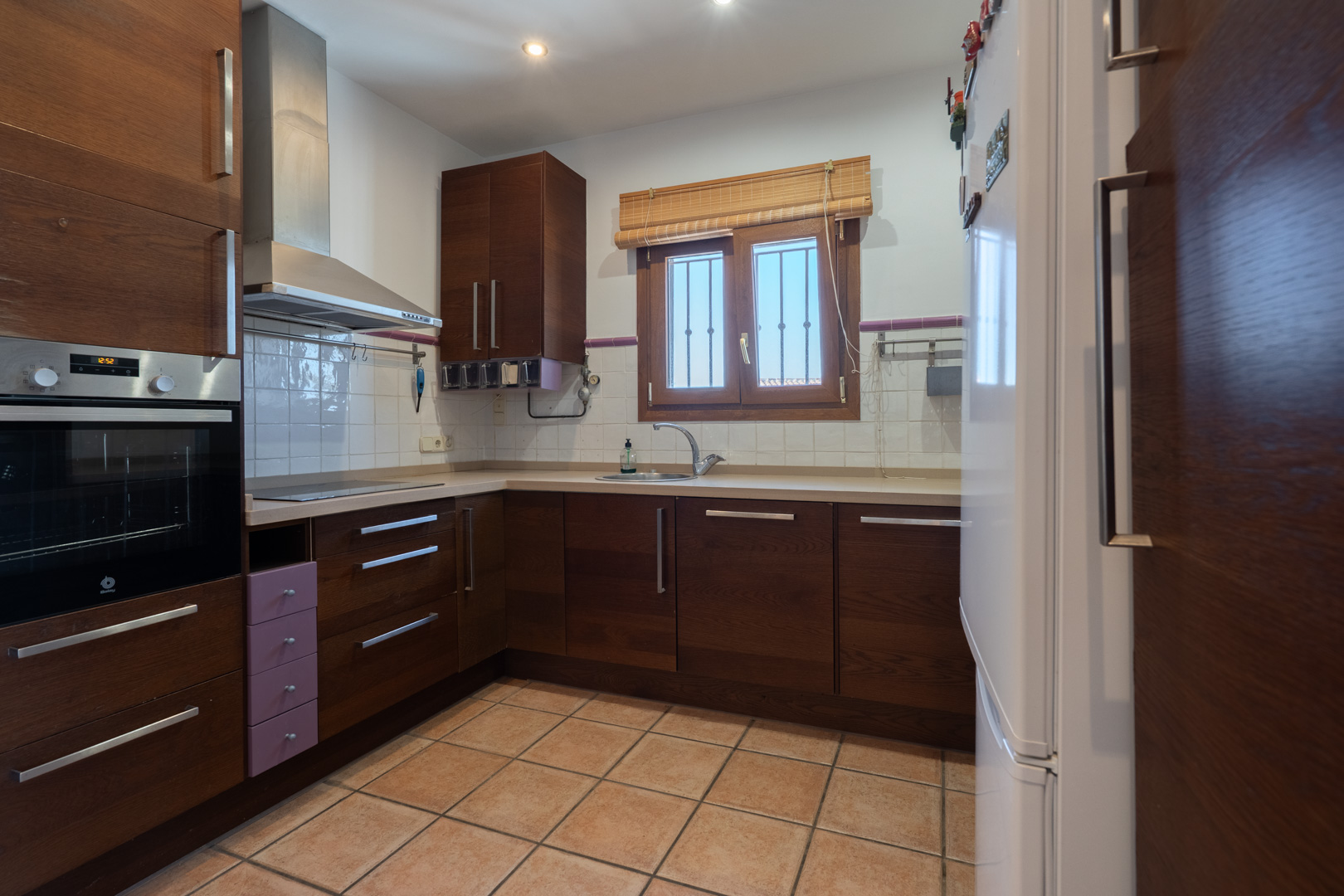 Cocina funcional con armarios de madera oscura, horno integrado Balay y suelo de gres. Incluye campana extractora de acero inoxidable y ventana con marco de madera.