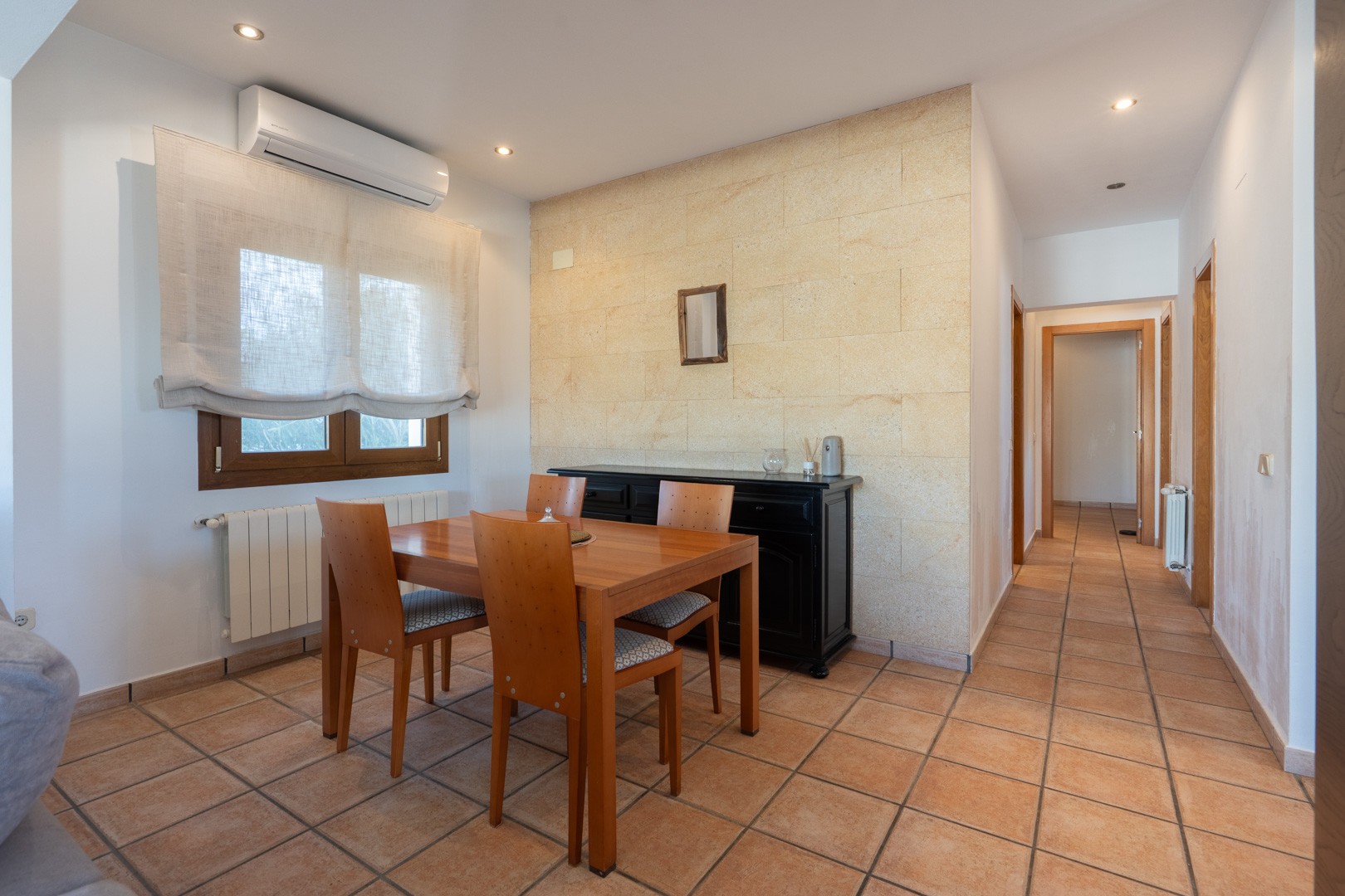 Comedor con pared de piedra natural, suelos de gres cerámico y mesa de madera. Incluye aire acondicionado, radiador y acceso al pasillo distribuidor.
