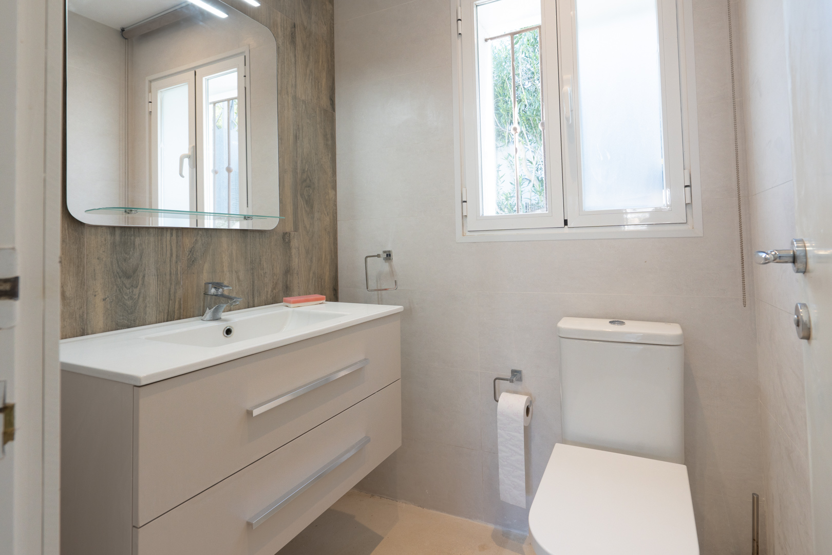 Cuarto de baño moderno con mueble de lavabo suspendido, pared de acento con efecto madera, espejo con repisa de cristal y ventana de PVC para luz natural.