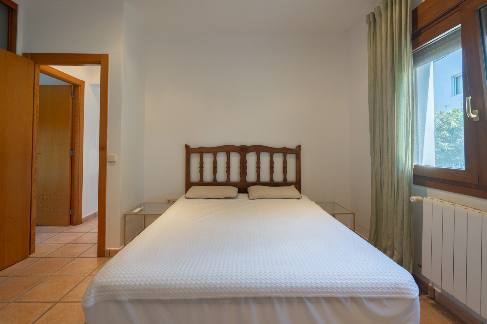 Dormitorio con cama doble, cabecero de madera tallada y suelos de gres. Incluye ventana con radiador, cortinas verdes y acceso al pasillo mediante puerta de madera.