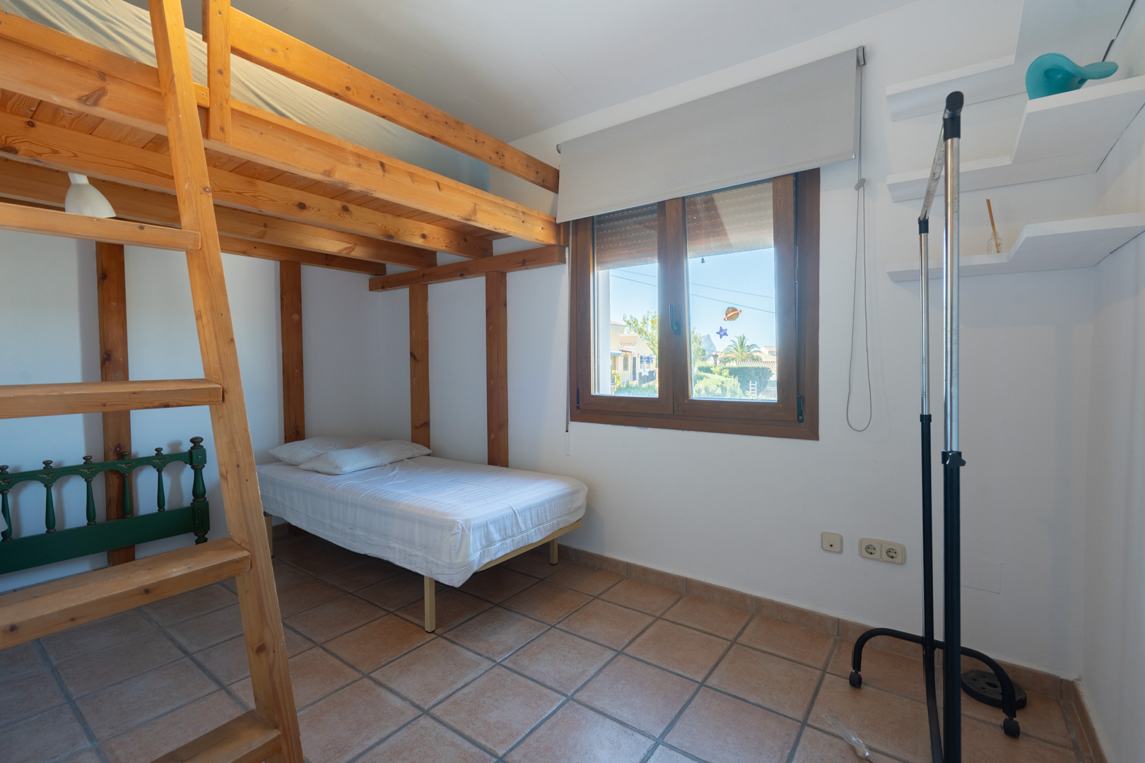 Dormitorio con estructura de litera de madera, suelos de gres rústico y ventana con marco de madera. Incluye estanterías blancas y luz natural.