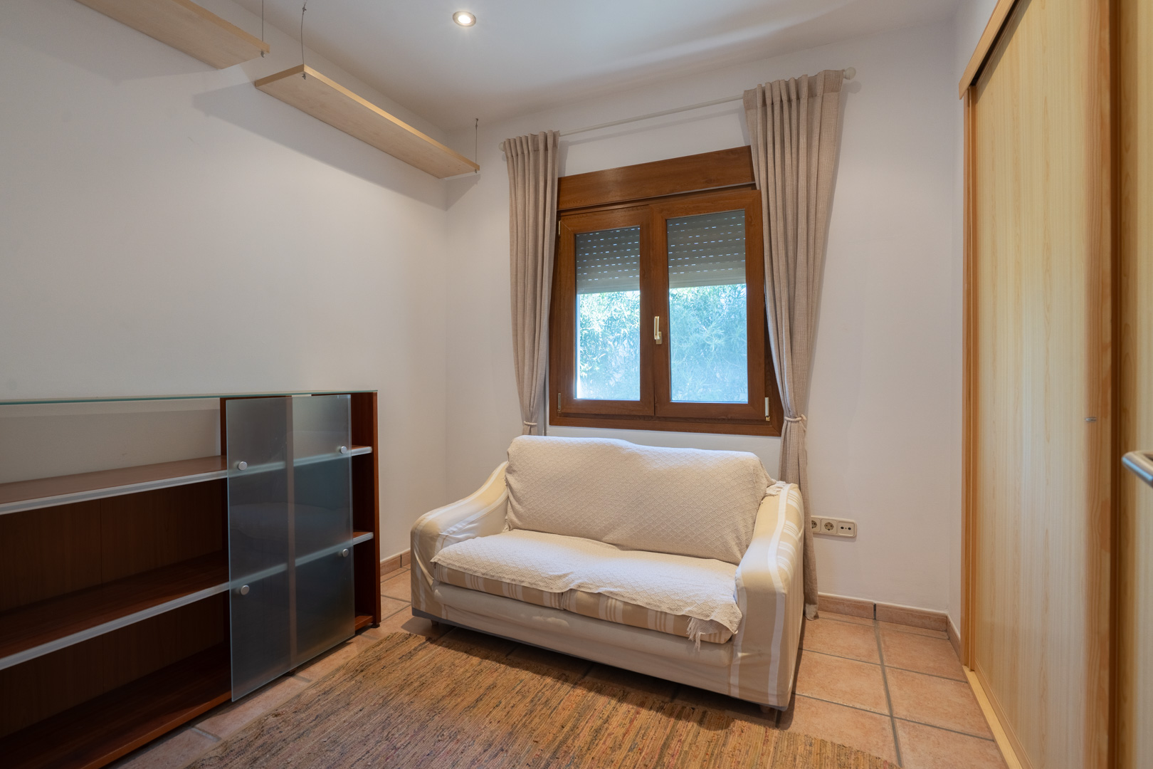 Dormitorio con suelos de gres, armario empotrado de madera clara y ventana de madera con persiana. Incluye estanterías y un sofá compacto.