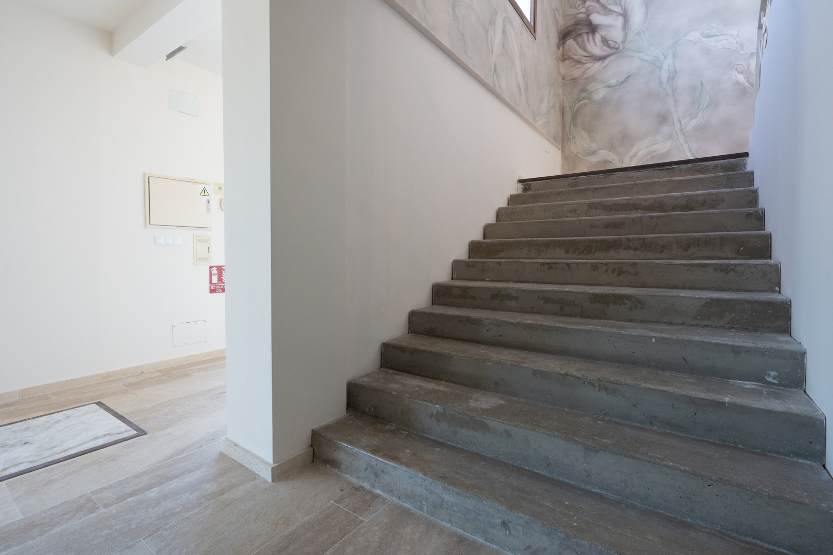 Escalera interior de hormigón visto que conecta los niveles, decorada con un mural floral en tonos suaves y suelos de gres porcelánico.