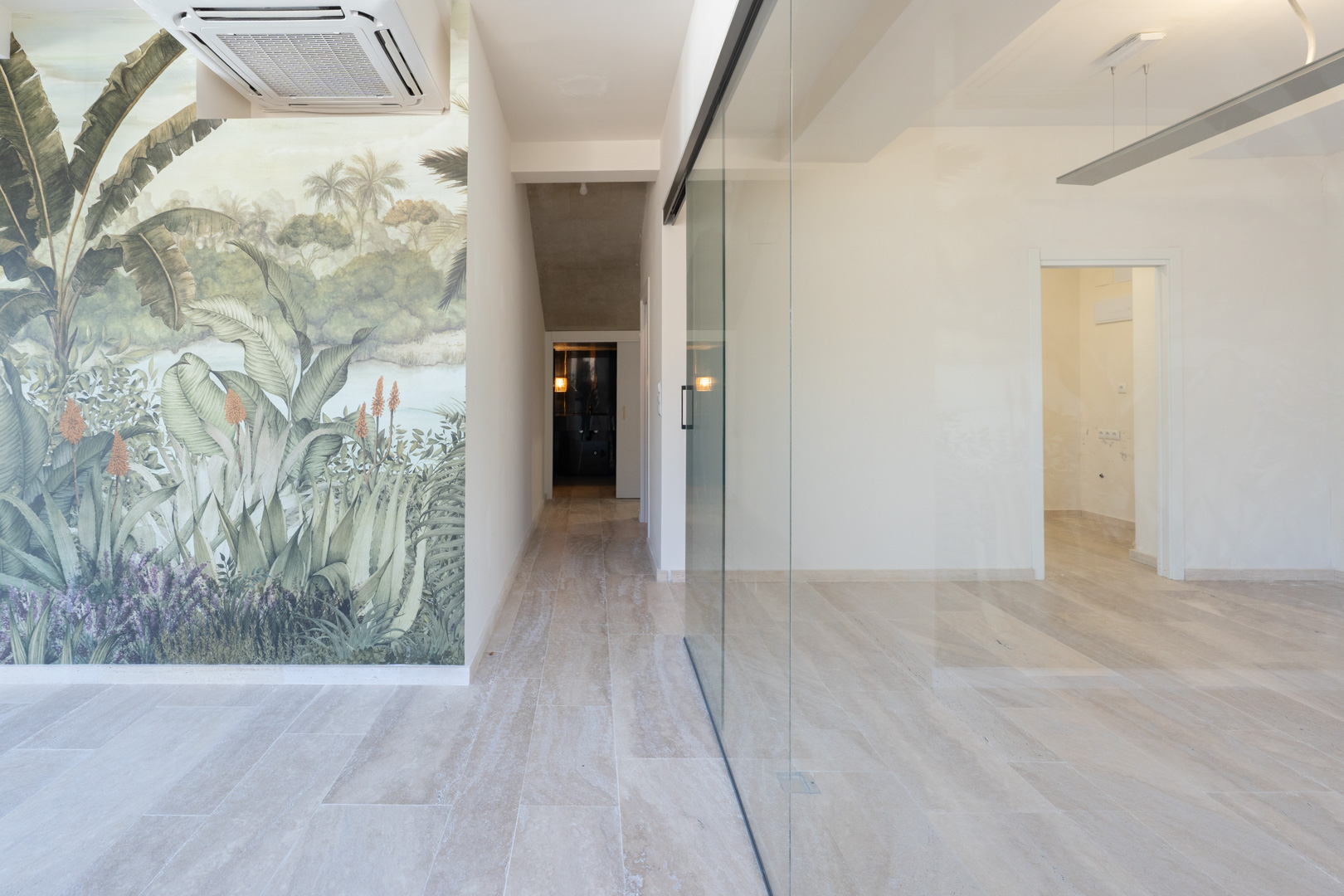 Espacio comercial diáfano con suelos de gres claro, un mural decorativo de temática tropical y una amplia división de cristal que separa las estancias.