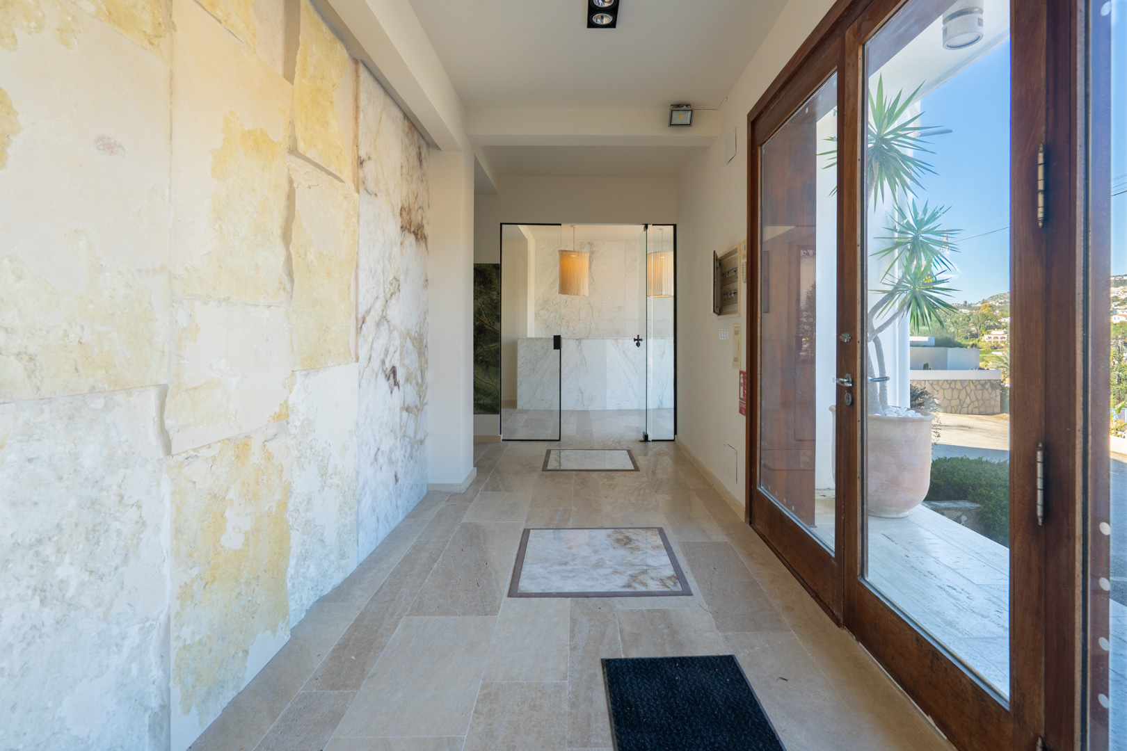 Hall de entrada con paredes de piedra natural, suelos de travertino con incrustaciones de mármol y acceso acristalado a la zona de recepción.