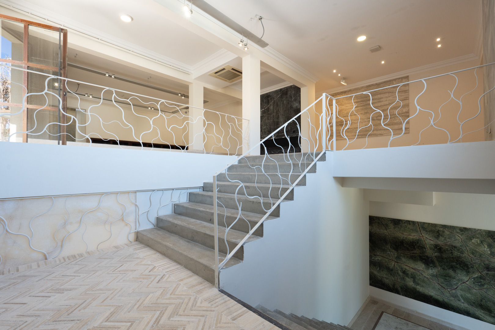 Impresionante escalera con barandilla de diseño orgánico que conecta los niveles comerciales, destacando suelos de espiga y paneles decorativos de piedra.