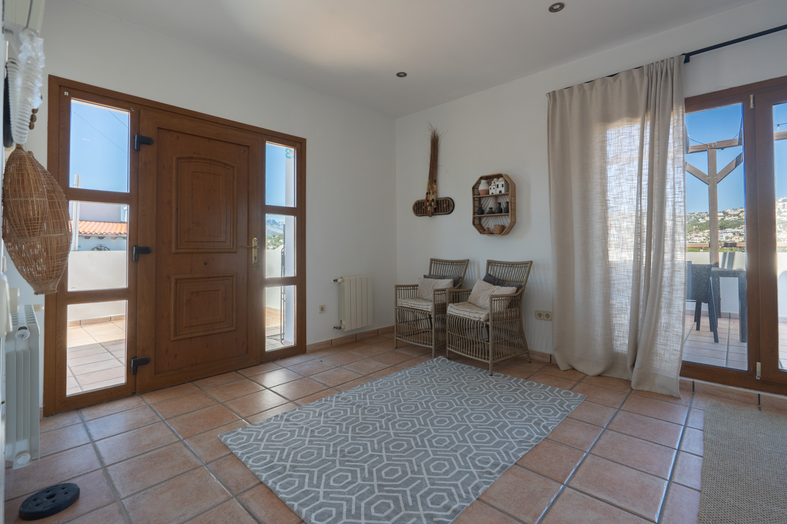 Recibidor luminoso con suelo de gres, puerta principal de madera con paneles acristalados y acceso a una terraza con pérgola de madera.