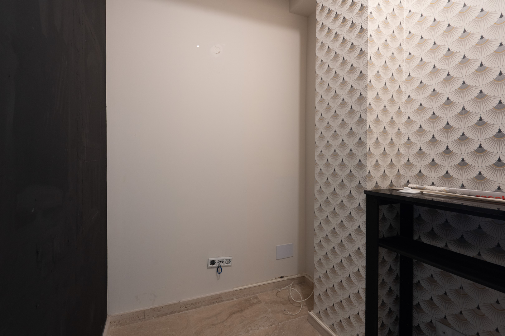 Rincón interior con pared decorada en papel pintado de diseño geométrico, suelo de piedra clara y tomas eléctricas integradas en la pared blanca.