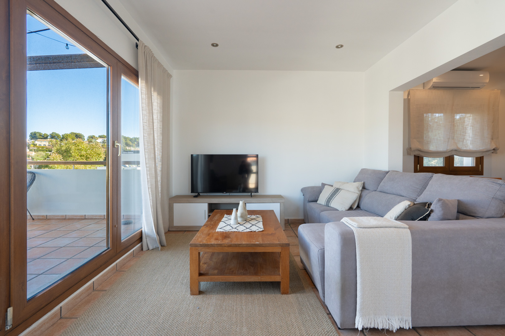 Salón luminoso con sofá gris, mesa de centro de madera y acceso directo a una terraza privada mediante grandes ventanales de madera.