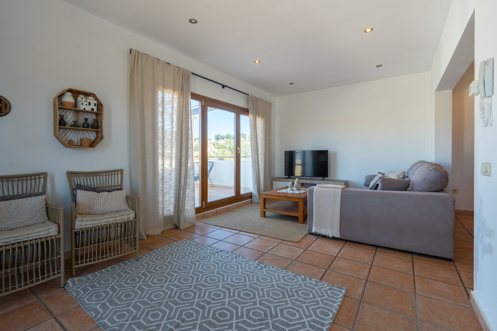 Salón luminoso con suelos de terracota, sofá rinconera gris y acceso directo a la terraza a través de amplios ventanales con marcos de madera.