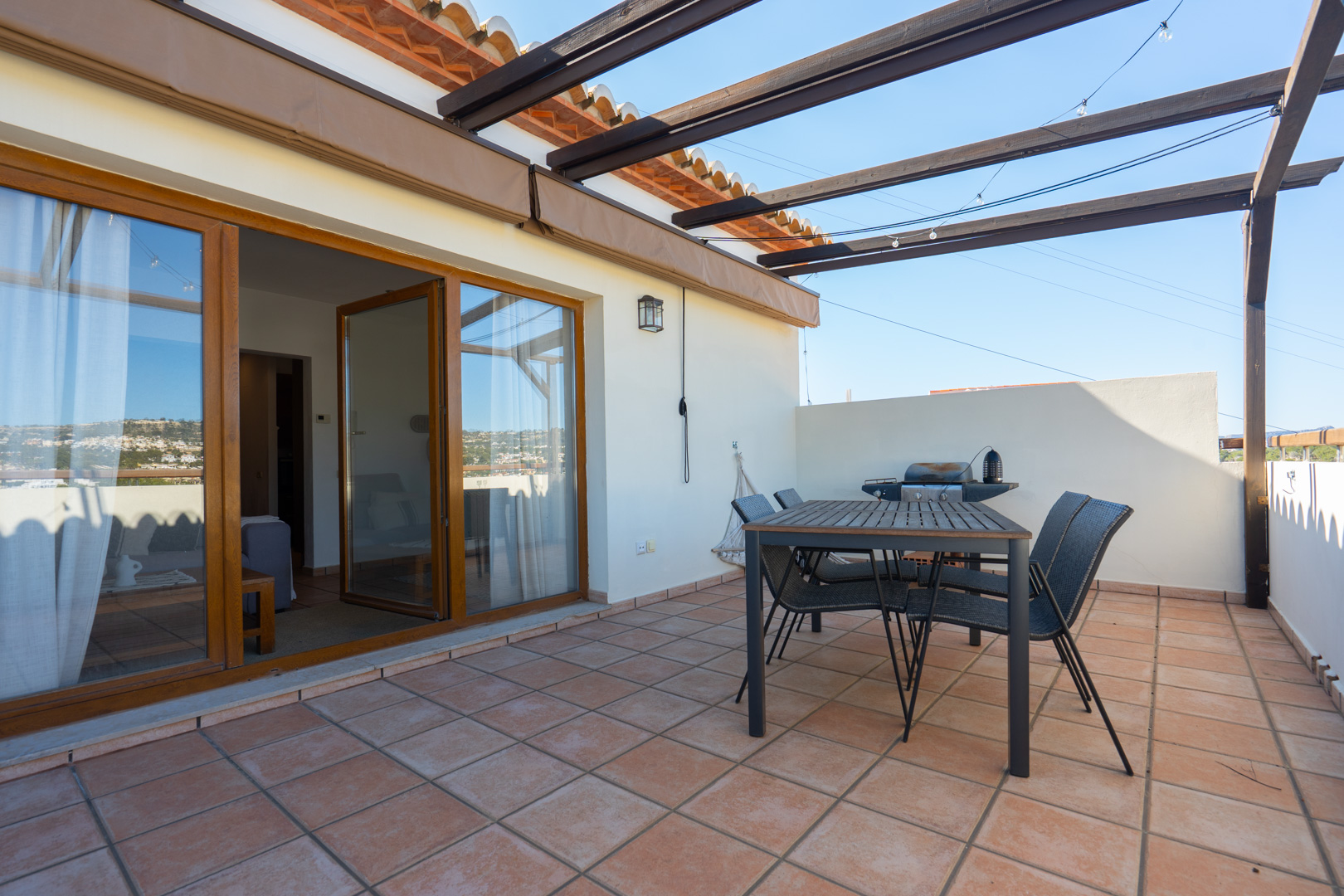 Terraza exterior con suelos cerámicos, zona de comedor con mesa de madera y pérgola. Incluye barbacoa y acceso directo al interior mediante amplios ventanales de madera.