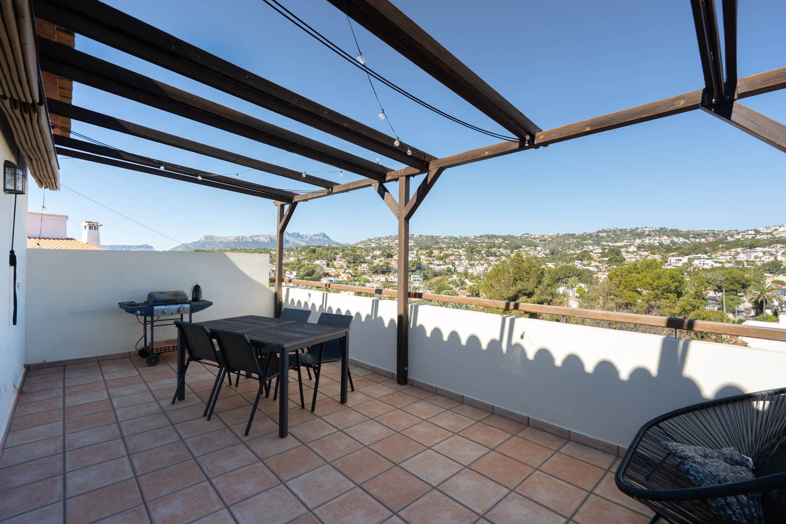 Terraza superior con suelo de terracota, pérgola de madera y zona de comedor. Ofrece vistas despejadas a la Sierra de Bernia y el entorno residencial de Moraira.