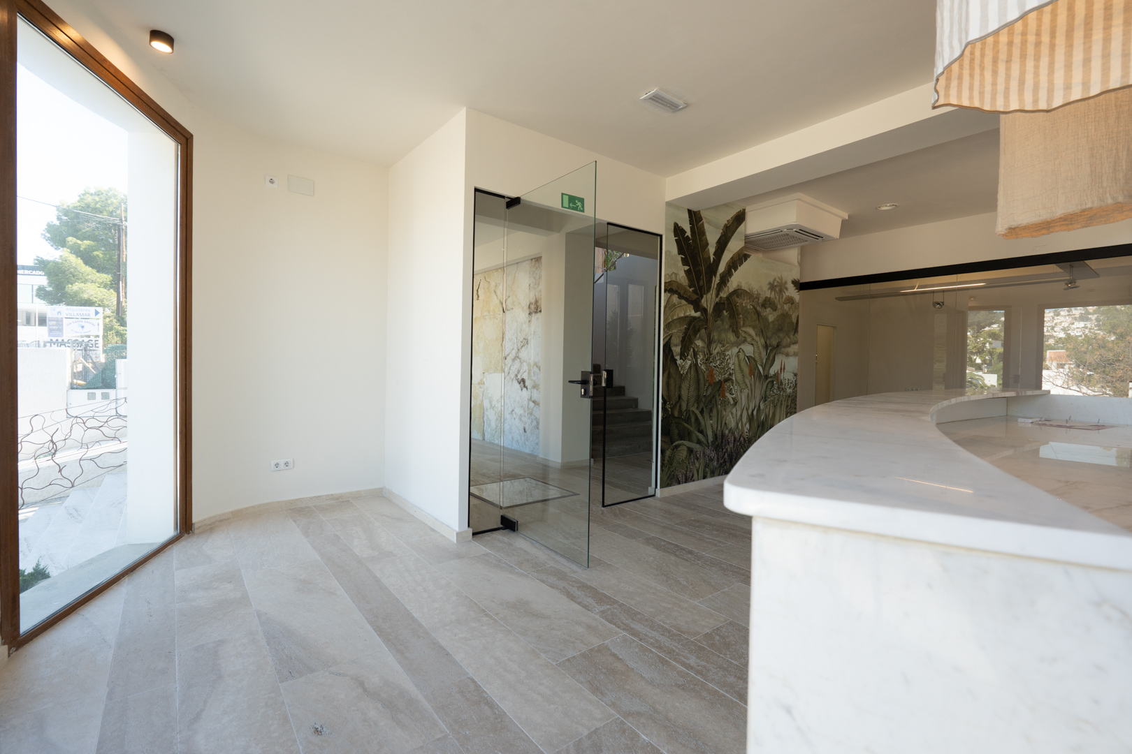 Zona de recepción con mostrador curvo de mármol blanco, suelos de piedra y puertas de cristal que dan paso a las estancias interiores y escaleras.