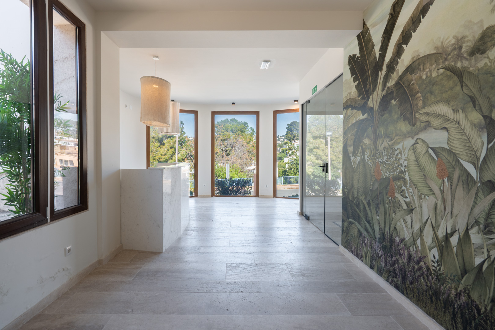 Zona de recepción con mostrador de mármol, suelos de piedra natural y mural botánico. Grandes ventanales con marcos de madera aportan luz natural y vistas al exterior.