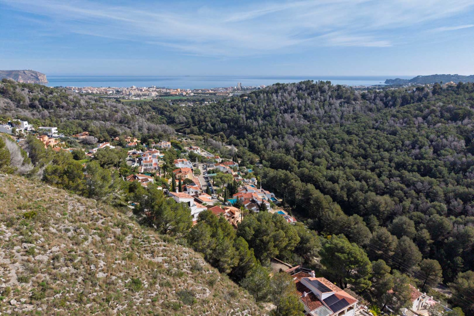 Vista aérea panorámica que muestra la topografía elevada de la zona sobre colinas boscosas, con vistas despejadas al mar Mediterráneo y la costa de Jávea.