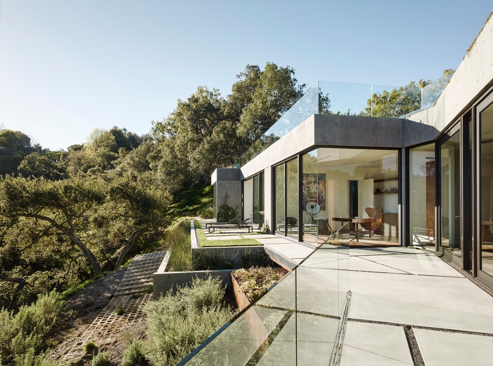 Exterior de villa moderna en hormigón con ventanales de suelo a techo y terraza con barandillas de cristal y pavimentos geométricos.