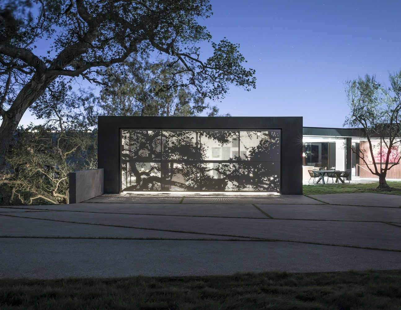 Fachada moderna con panel metálico perforado retroiluminado que proyecta motivos de árboles, integrada en un diseño minimalista con pavimentos geométricos y zona de comedor exterior.