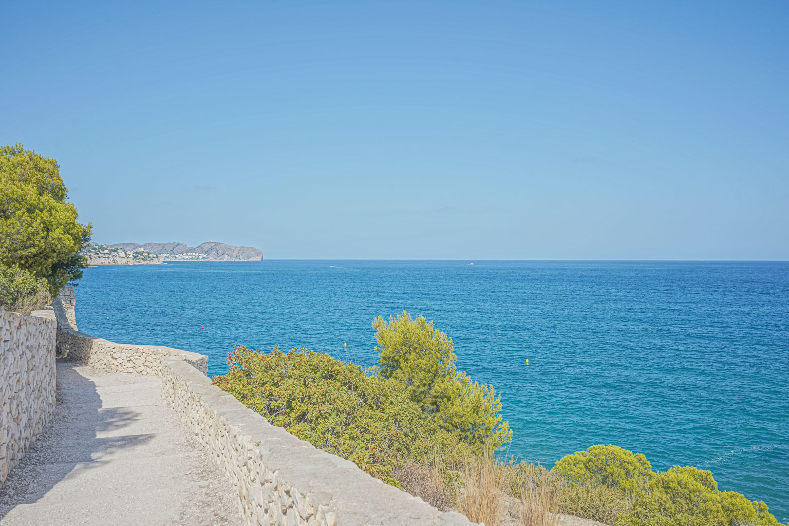 Paseo costero con muros de piedra seca y vistas panorámicas al mar Mediterráneo, rodeado de vegetación autóctona y pinos bajo un cielo despejado.