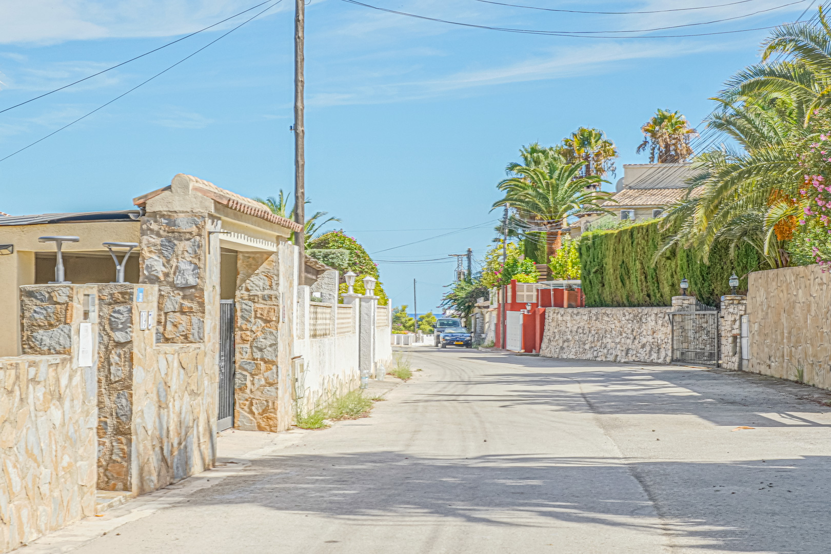 Vista de la calle residencial de acceso, flanqueada por muros de piedra natural y vegetación mediterránea, con el mar visible al fondo bajo un cielo despejado.