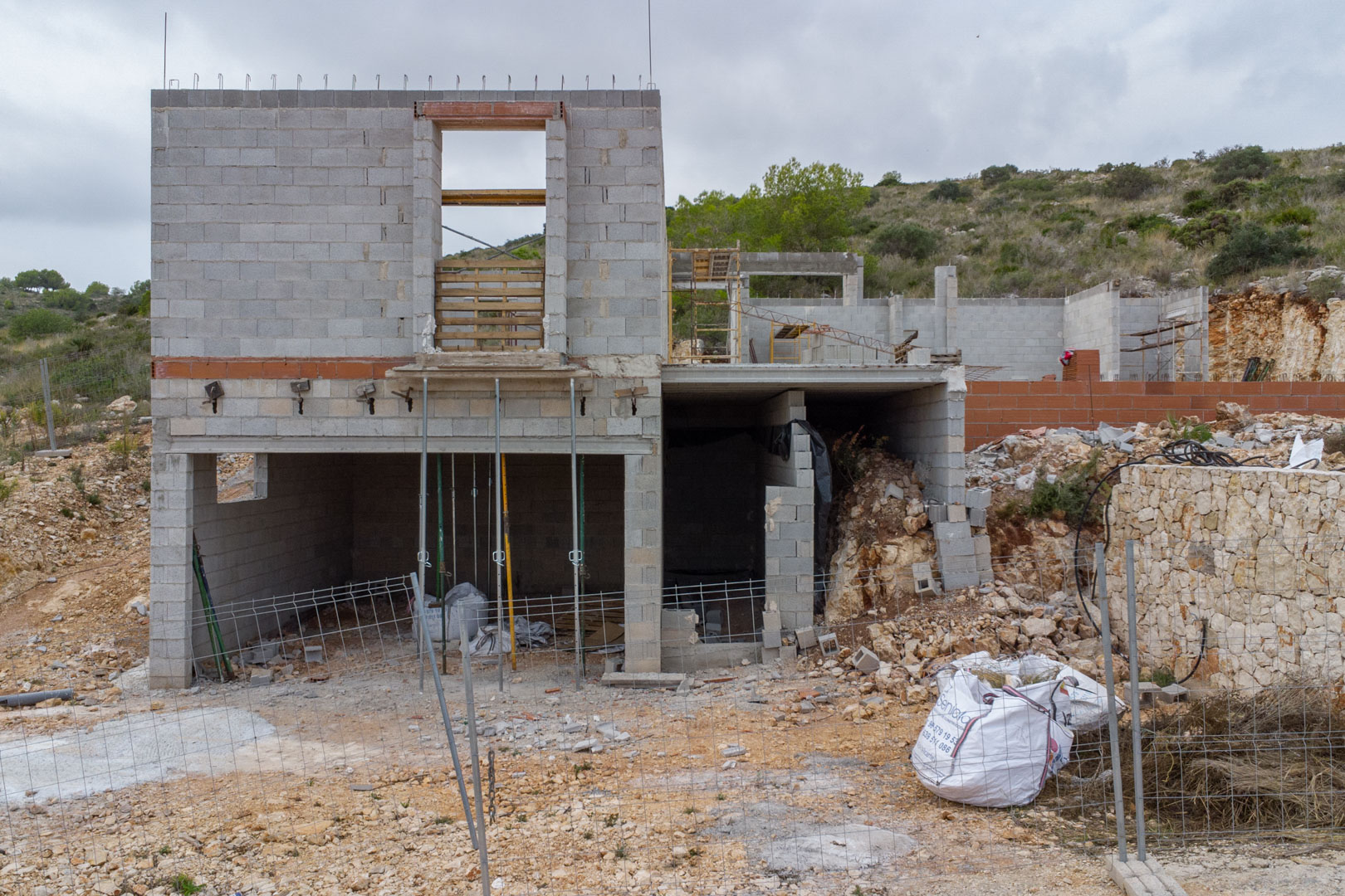 Vista exterior de la estructura de bloques de hormigón en construcción, con varios niveles y un muro de contención de piedra natural en una ladera de Pedreguer.
