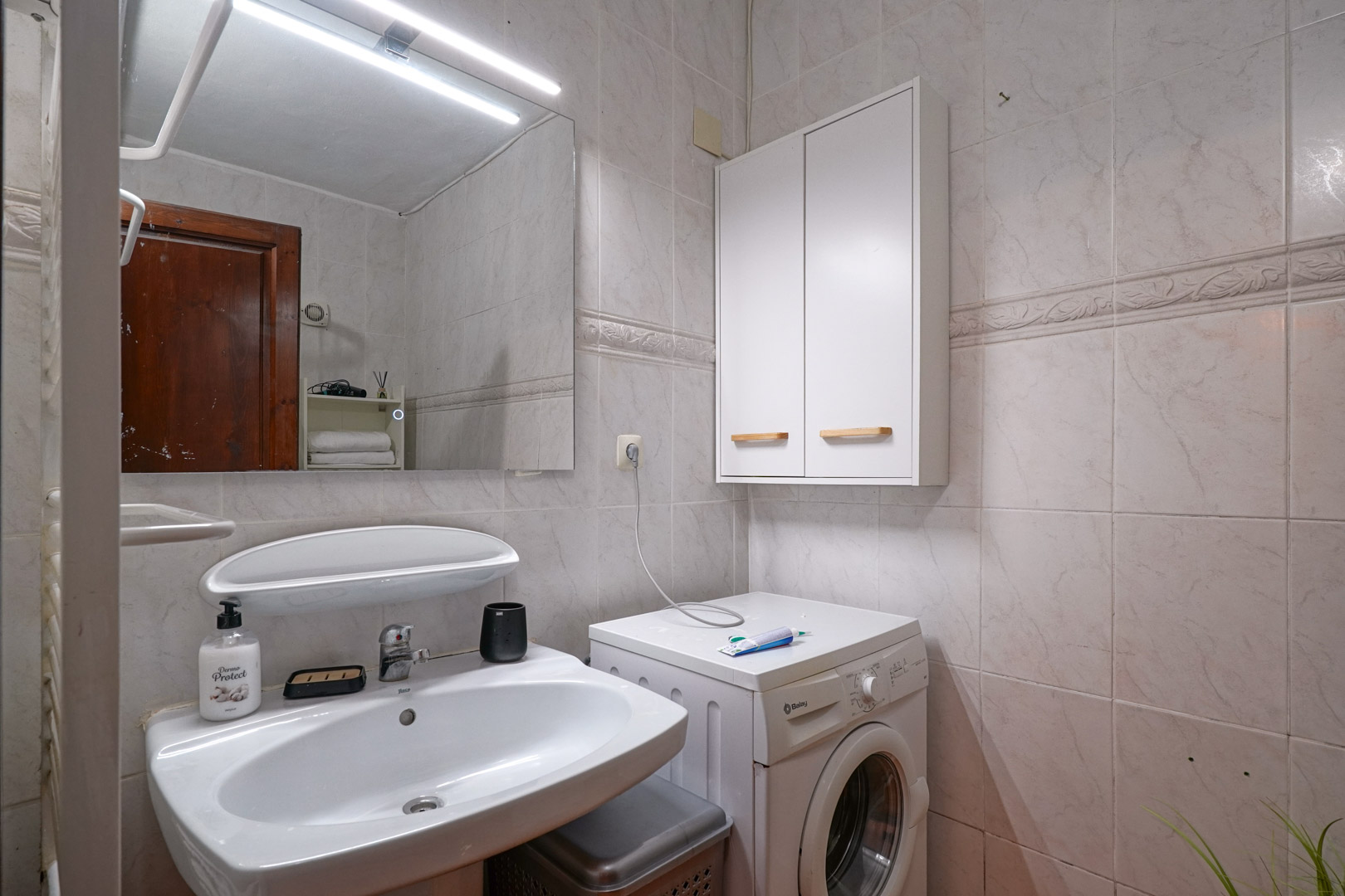 Baño funcional con lavabo cerámico, espejo con iluminación LED superior y zona de lavandería integrada con lavadora y armario auxiliar sobre paredes alicatadas.
