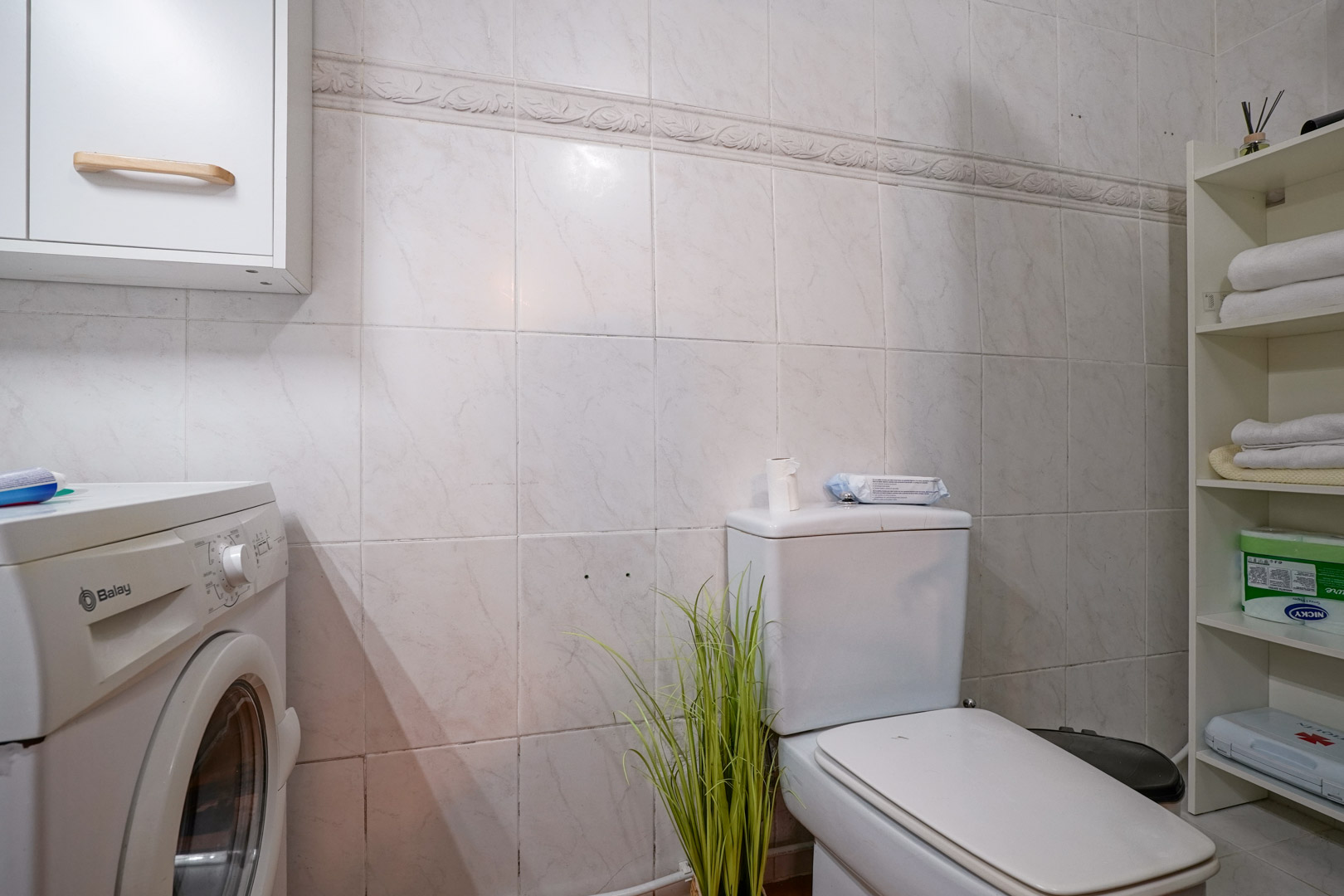 Baño funcional con zona de lavandería, equipado con lavadora Balay, estanterías de almacenaje y paredes alicatadas con cenefa decorativa.