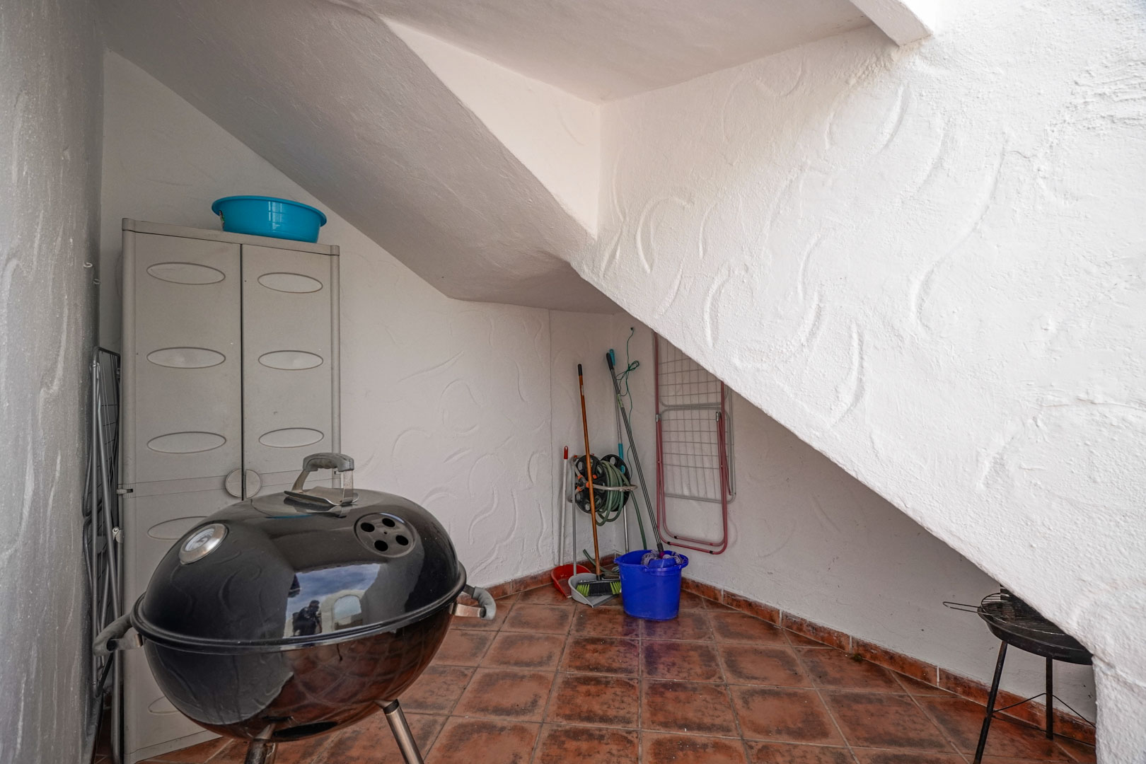 Espacio de almacenamiento exterior bajo la escalera con suelos de terracota, armario de resina y zona para barbacoa y utensilios de limpieza.