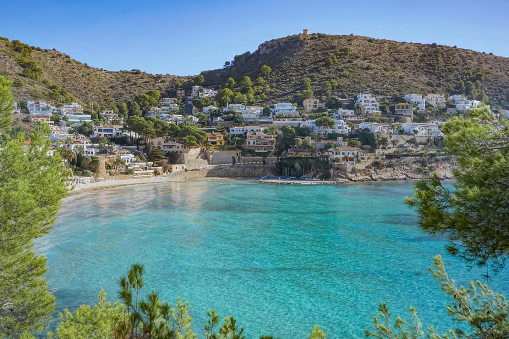Vista panorámica de la cala de El Portet en Moraira, con sus aguas turquesas y villas integradas en la ladera bajo la histórica torre vigía del Cap d'Or.