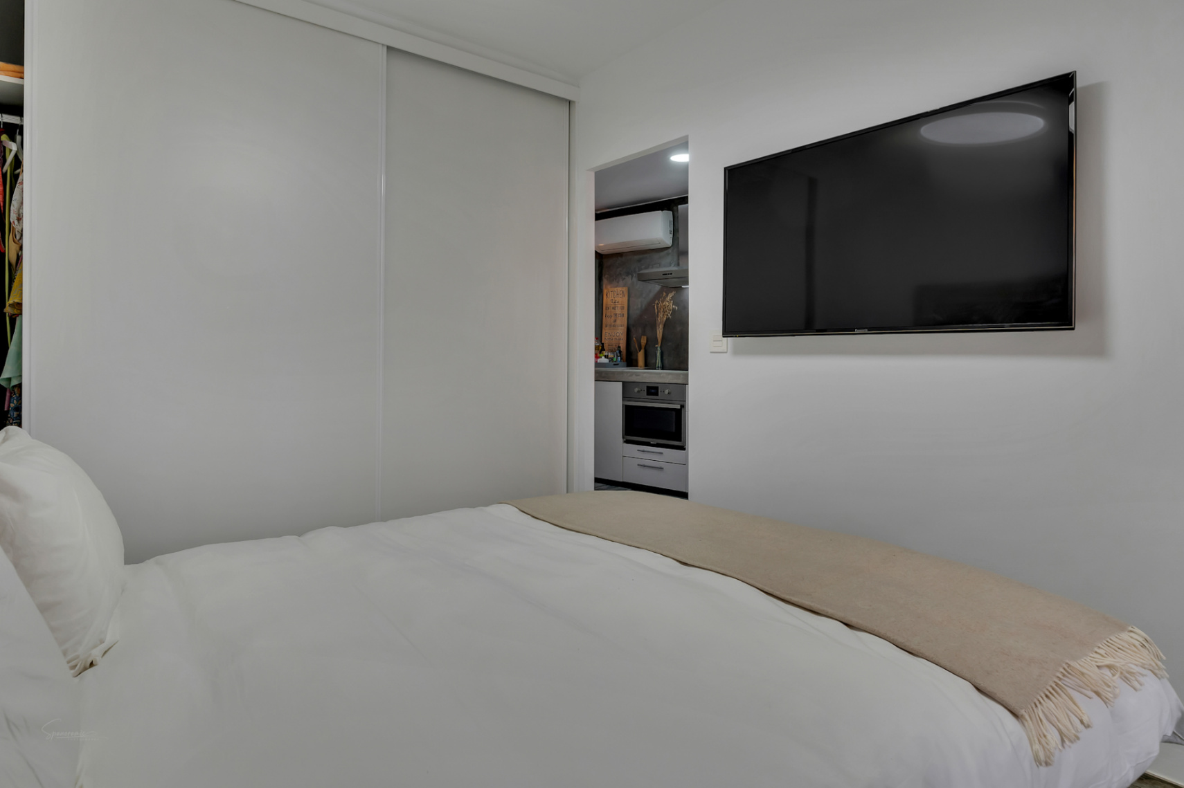 Dormitorio moderno con cama blanca y TV de pantalla plana. Cocina compacta visible al fondo.
