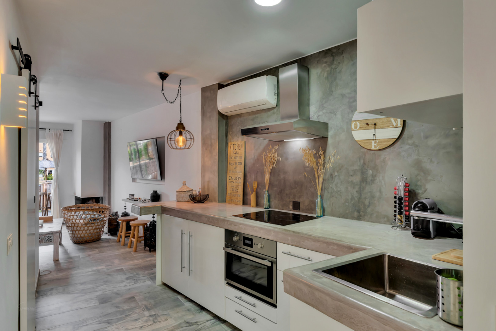 Vista de la cocina moderna con encimeras de microcemento y pared decorativa. Se aprecia el salón con chimenea al fondo.